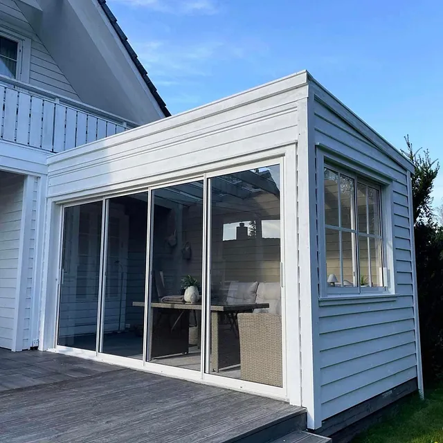 Glassinnbygget tilbygg med hvit kledning, skyvedører og tilhørende terrasse mot en grønn plen.