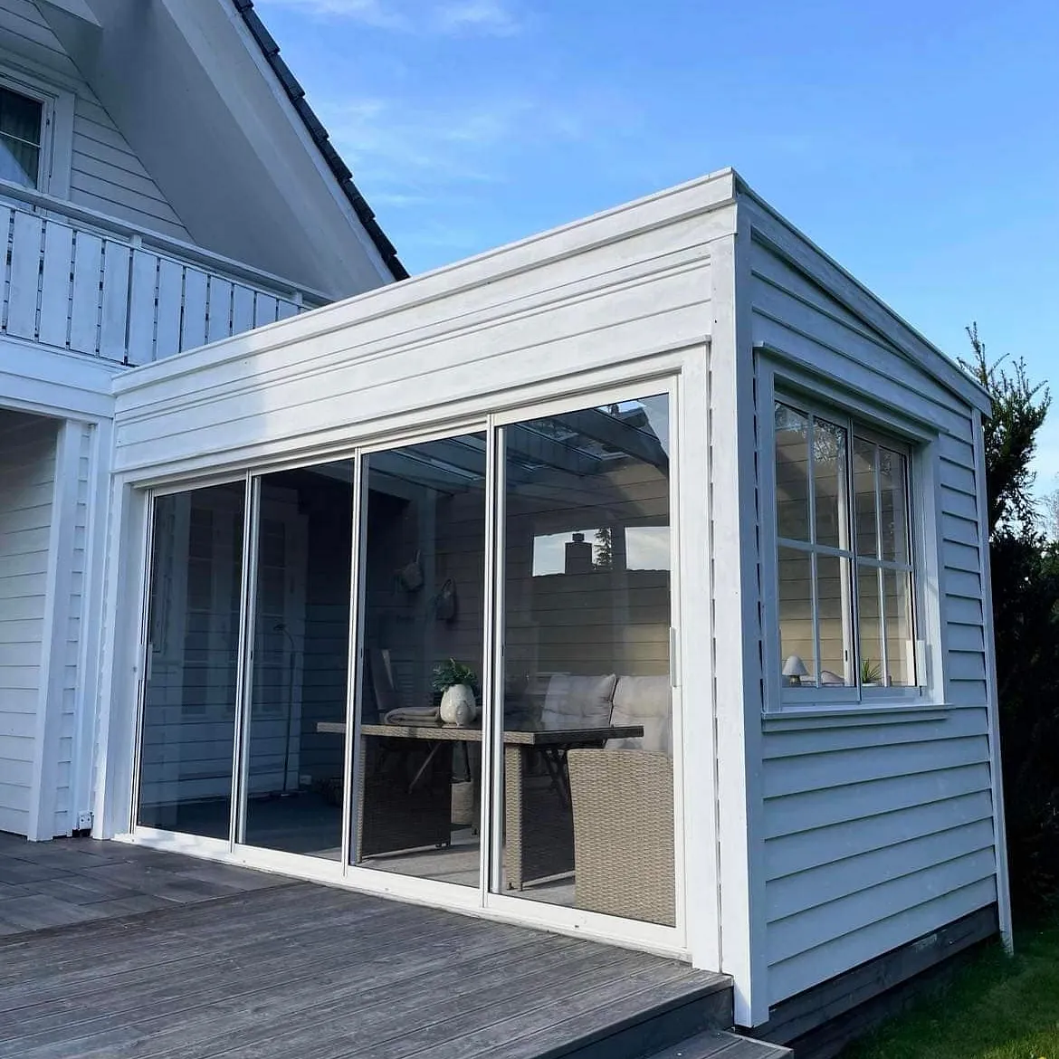 Glassinnbygget tilbygg med hvit kledning, skyvedører og tilhørende terrasse mot en grønn plen.