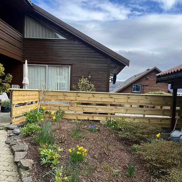Hage med blomsterbed og lavt tregjerde foran et hus med mørk trekledning og store vinduer.