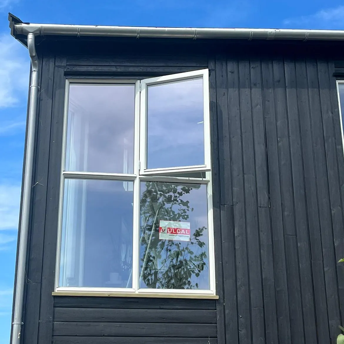 Moderne svart fasade med stort vindu fra Uldal, der øvre del står på gløtt under blå himmel.